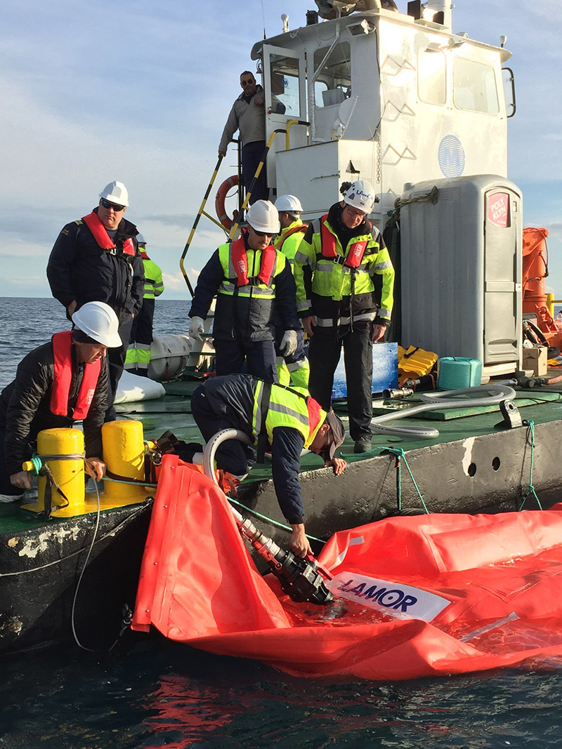 Des intervenants en position sur une barge pour déployer une citerne remorquable en tissu.
